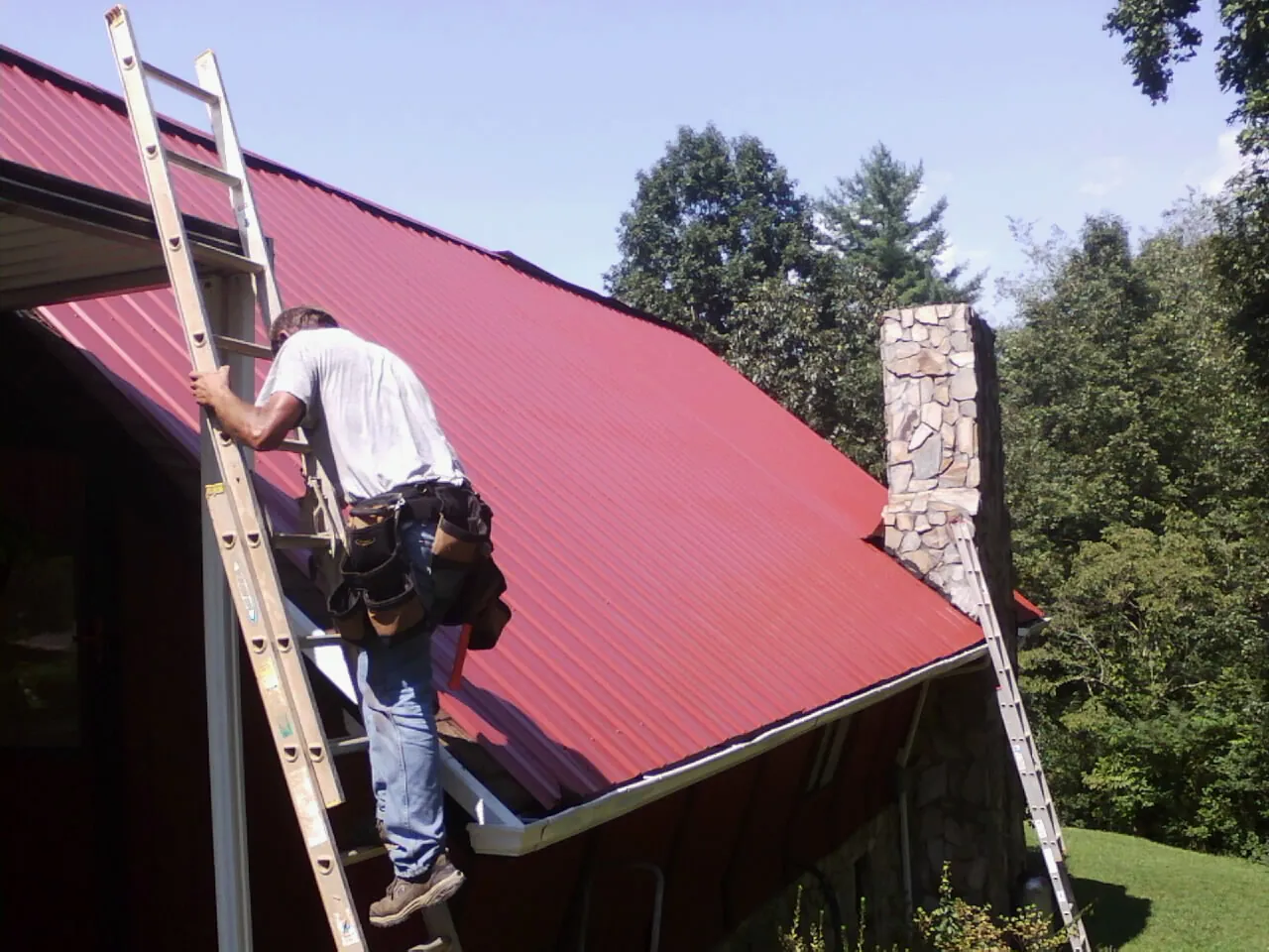 Roofer on a ladder beside a red metal roof and stone chimney — Soffit & Fascia Repair by Roofing Brothers in Davenport Center